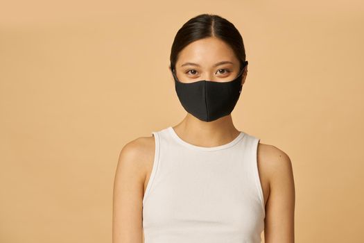 Beautiful young woman wearing black facial mask looking at camera while posing isolated over beige background. Safety, pandemic concept