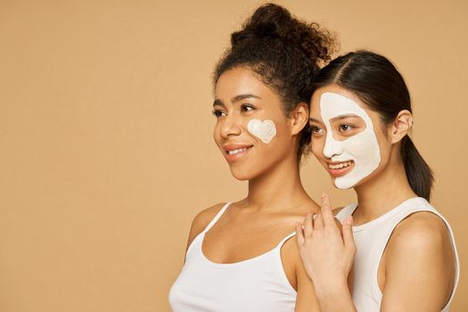 Portrait of two young women, female friends smiling aside while posing together with facial masks on isolated over beige background. Skincare, beauty concept