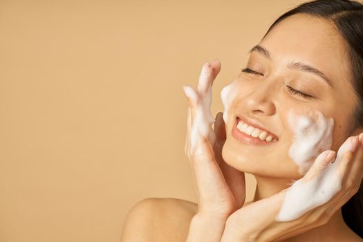 Beauty portrait of lovely young woman smiling with eyes closed while applying gentle foam facial cleanser isolated over beige background. Beauty products and skin care concept