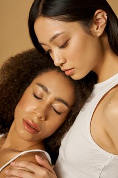 Face closeup of two beautiful mixed race young women with perfect glowing skin posing isolated over light brown background. Female beauty portrait. Selective focus