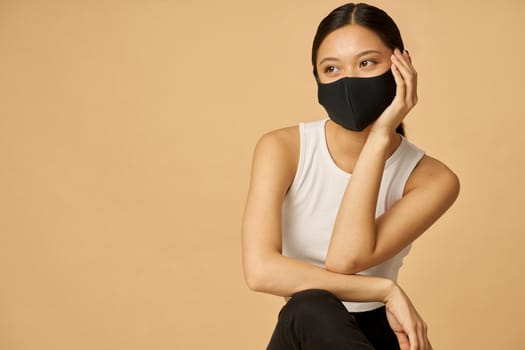 Dreamy young mixed race woman wearing black facial mask, staying safe, looking aside while posing isolated over beige background. Safety, pandemic concept