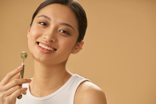 Portrait of joyful young woman smiling at camera, using jade roller for massaging her face, posing isolated over beige background. Skincare concept