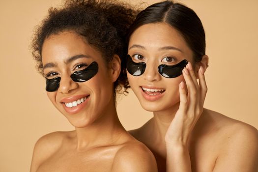 Beauty portrait of two happy young women posing with applied black under eye patches isolated over beige background. Skincare routine concept