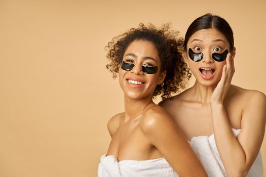 Two excited young women wrapped in towels posing with applied black hydro gel under eye patches on facial skin isolated over beige background. Skincare routine concept