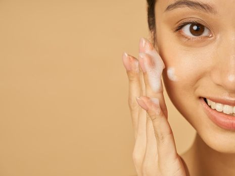 Studio half face portrait of beautiful young woman smiling at camera while applying gentle foam facial cleanser isolated over beige background. Beauty products and skin care concept