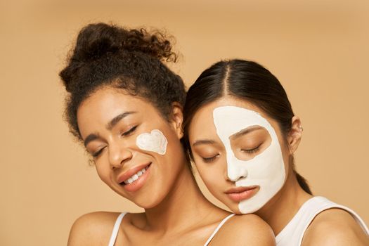 Close up portrait of two young female friends with facial masks on posing with eyes closed isolated over beige background. Skincare, beauty concept