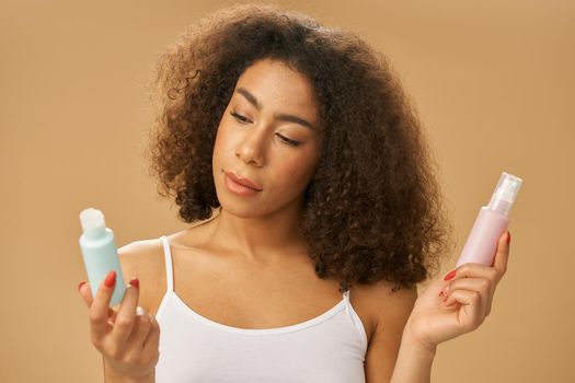 Portrait of attractive healthy african american young woman choosing between two bottles with beauty product while posing over beige background. Skincare concept