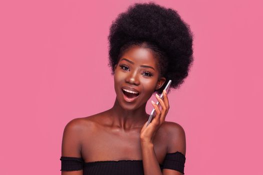 Amusing African American woman chatting with girlfriends on phone or smartphone. Female model with afro hairstyle smiling in black cut top with bared shoulders isolated on pink background.