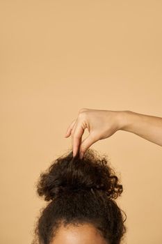 Studio shot of young mixed race woman with highly raised dark curly hair fixing her hairstyle, posing isolated over beige background. Vertical shot