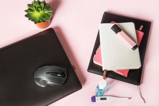 Top view of the desktop with notebooks, marker, laptop, thermometer and hand wash gel. Work from home using an online device, protection from coronavirus during the quarantine period. Pink background.