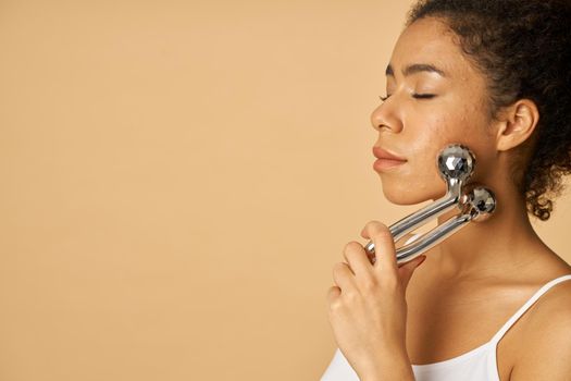 Relaxed young woman posing with eyes closed while using silver metal face roller isolated over beige background. Facial massage concept