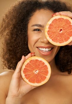 Portrait of mixed race young woman looking happy while posing with grapefruit cut in half over beige background. Health and beauty concept. Front view