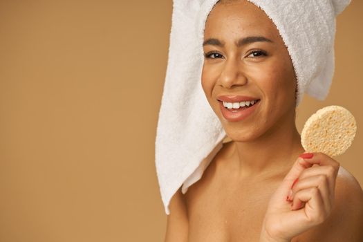 Joyful mixed race young woman smiling at camera, holding cleansing face sponge, posing isolated over beige background. Bathroom routine. Youth and skin care concept. Horizontal shot