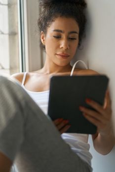 Relaxed young woman reading a book using tablet pc while sitting by the window at home. Lifestyle, leisure concept