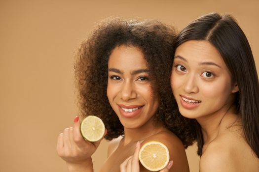 Two cute mixed race young women looking at camera and holding lemon and lime cut in half while posing together isolated over beige background. Health and beauty concept