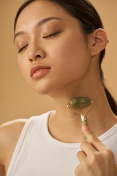 Portrait of young woman looking relaxed while using jade roller for massaging her neck, posing isolated over beige background. Skincare concept. Vertical shot