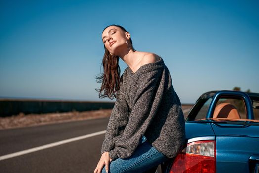 Young woman near roofless car and smiling outdoors on sunny day near the empty road. No people, sportive blue car