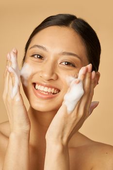 Beauty portrait of joyful young woman smiling at camera while applying gentle foam facial cleanser isolated over beige background. Beauty products and skin care concept