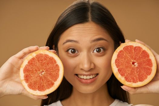 Portrait of mixed race young woman looking excited at camera, holding grapefruit cut in half while posing isolated over beige background. Health and beauty concept. Front view