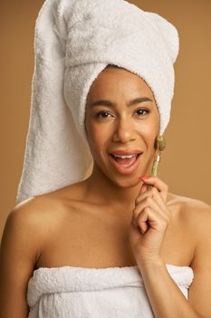 Playful mixed race young woman after shower looking at camera while using jade roller for massaging her face isolated over beige background. Skincare concept