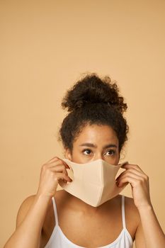 Portrait of adorable young mixed race woman putting on protective facial mask, posing isolated over beige background. Safety, pandemic concept