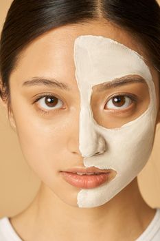 Close up portrait of young woman with facial mask applied on half of her face receiving spa treatments, posing isolated over beige background. Front view