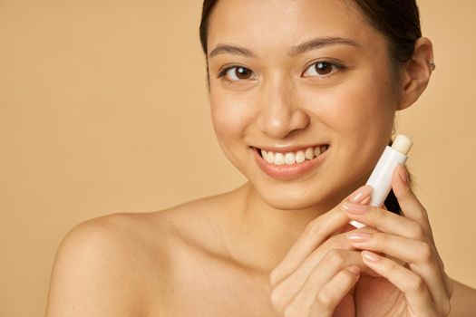 Close up portrait of lovely young woman smiling while holding lip balm, posing isolated over beige background. Beauty treatment concept