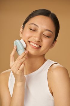 Beautiful young woman looking happy while using silicone facial cleansing brush, posing isolated over beige background. Skincare concept