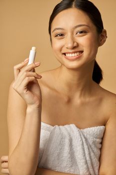 Cheerful natural young woman wrapped in towel smiling at camera, holding lip balm while posing isolated over beige background. Beauty treatment concept