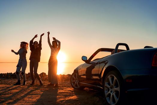 Cheerful young three women are dancing near cabriolet. Beautiful sunset at the background