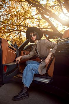 Sexy lady in the roofless car. Stylish woman is wearing sunglasses and black hat. She had fashion haircut. Autumn season.