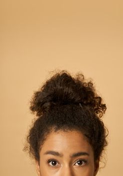 Cropped portrait of young mixed race woman with highly raised dark curly hair looking at camera while posing isolated over beige background. Vertical shot