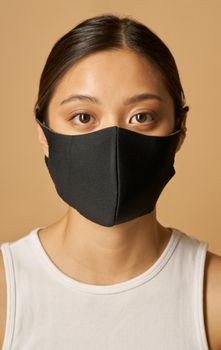 Studio portrait of beautiful young mixed race woman wearing black facial mask, looking at camera while posing isolated over beige background. Safety, pandemic concept