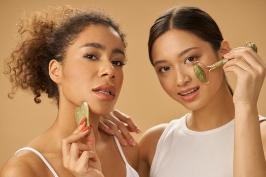 Beauty treatment. Attractive young women looking at camera, using jade roller and facial gua sha while posing together isolated over beige background. Skincare concept