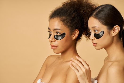 Beauty portrait of two happy young women looking away, posing with applied black hydro gel under eye patches isolated over beige background. Skincare routine concept