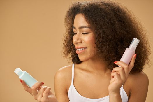 Attractive young woman with curly hair looking cheerful while choosing between two beauty products, posing isolated over beige background. Skincare concept