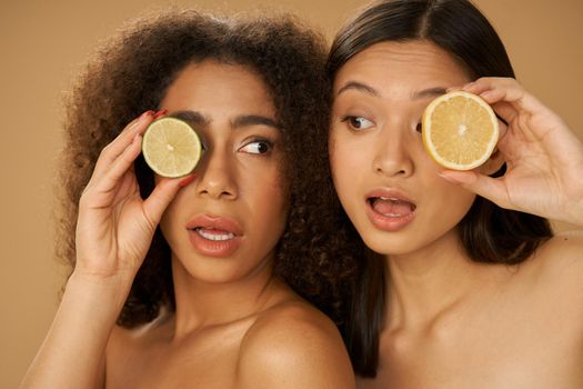 Portrait of two funny mixed race young women looking surprised, holding lemon and lime cut in half while posing together isolated over beige background. Health and beauty concept