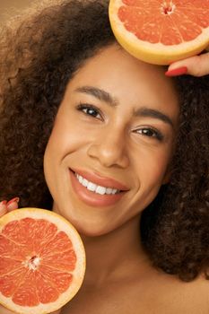 Portrait of cheerful mixed race young woman smiling at camera, holding grapefruit cut in half while posing over beige background. Health and beauty concept. Front view