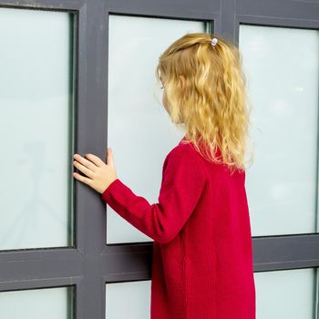 Rear view of a girl looking at a wall or window, charming blond girl in a red knitted dress portrait of a cute preschooler child. Close-up