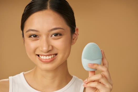 Cute young woman smiling at camera while holding silicone facial cleansing brush, posing isolated over beige background. Washing accessory. Skincare concept