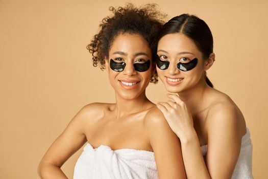 Portrait of two cheerful young women wrapped in white towel posing with applied black under eye patches, standing isolated over beige background. Beauty treatment concept