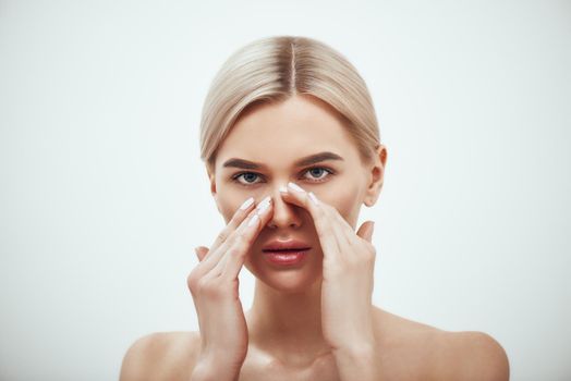 Rhinoplasty - nose surgery. Portrait of attractive blonde woman touching her nose and looking at camera while standing against grey background. Plastic surgery concept. Healthcare