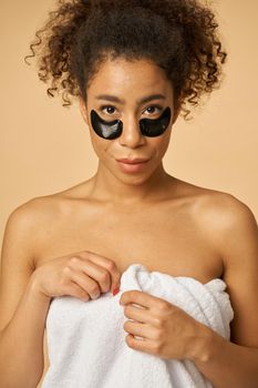 Portrait of young woman wrapped in white towel after shower posing with applied black under eye patches isolated over beige background. Bathroom routine concept