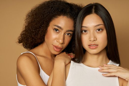 Beauty portrait of two mixed race young women with perfect skin looking at camera while posing together, standing isolated over light brown background. Skincare, diversity concept