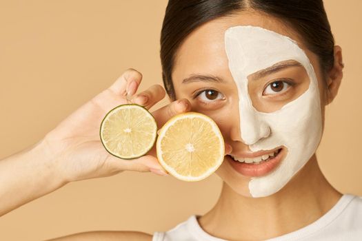 Young woman with facial mask applied on half of her face holding slice of lime and lemon, posing isolated over beige background. Skincare, natural cosmetics concept