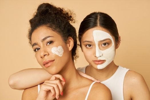 Portrait of two young women, female friends looking at camera while posing together with facial masks on isolated over beige background. Skincare, beauty concept