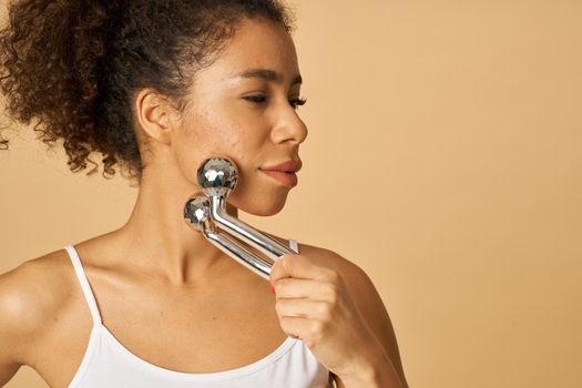 Portrait of attractive young woman looking aside while using silver metal face roller, posing isolated over beige background. Facial massage concept