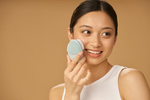 Lovely young woman smiling aside while using silicone facial cleansing brush, posing isolated over beige background. Skincare concept