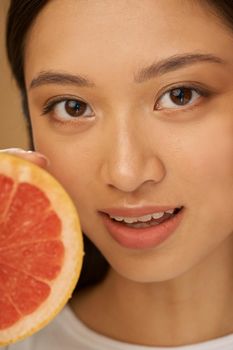 Close up portrait of attractive mixed race young woman looking at camera, holding grapefruit cut in half, posing isolated over beige background. Health and beauty concept