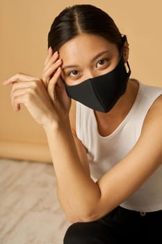 Portrait of adorable young mixed race woman wearing black facial mask, staying safe while posing for camera isolated over beige background. Safety, pandemic concept